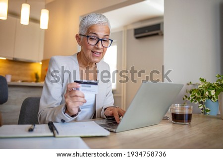 Happy senior woman making online payments of bill using laptop. Smiling mature woman shopping online with credit card. Pensioner holding credit card for internet banking and looking at camera.