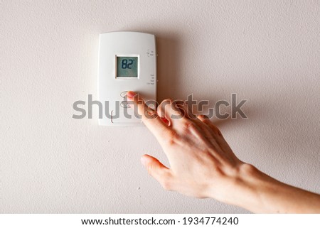 A woman is pressing the up button of a wall attached house thermostat with digital display showing the temperature. A concept image for electricity bill, heating, cooling, eco friendly, saving etc. Royalty-Free Stock Photo #1934774240