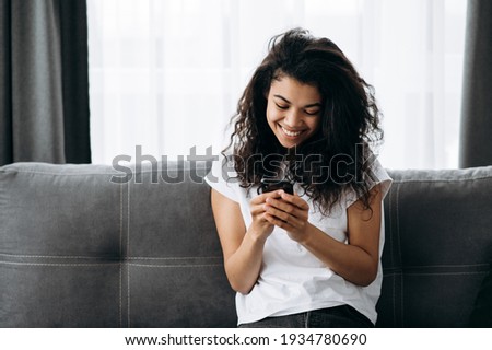 African american pretty woman sit at the couch in living room and use smartphone. Beautiful young woman in casual wear is chatting with friends or family, browsing internet, and smiling