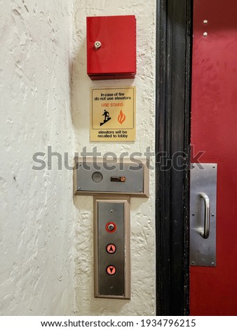 An elevator has an old fashioned steel door, painted red, covering the elevator doors. Beside this are call buttons, a locked fire box, and instructions in case of fire.