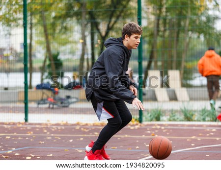 Cute Teenager with orange basketball ball plays basketball on street playground in spring summer. Hobby, active lifestyle, sports activity for kids.