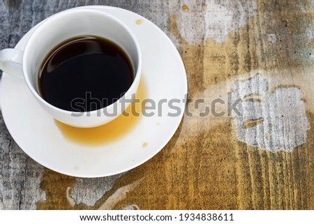 A white cup sits on a saucer in a puddle of spilled coffee on a wooden table.