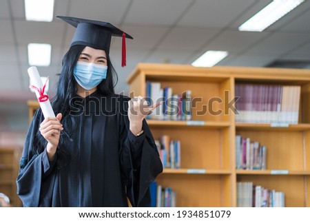 A young cheerful Asian woman university graduate in graduation gown and cap wears a sanitary mask holds a degree certificate to celebrate her education achievement on the commencement day. Stock photo