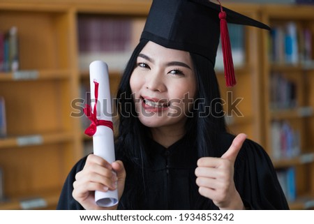 A young happy Asian woman university graduates in graduation gown and cap wears a face mask holds a degree certificate to celebrate her education achievement on the commencement day. Stock photo