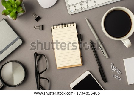 Overhead photo of notebook keyboard computer mouse magnifier pens phone plant glasses cup of coffee and paperclips isolated on the grey background