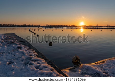 A beautiful sunset seen from a snowy boat dock where a line with buoys runs to an island with resting cormorants in lake Zoetermeerse Plas