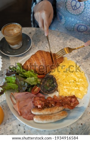 Woman cuts croissant that prepared with scramble egg, ham, bacon, sausages, salad and truffle mushroom in white dish. Breakfast and Coffee picture