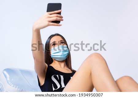 A brunette girl in a swimsuit and a medical mask on an inflatable mattress makes a selfie on a smartphone. Rest during a pandemic on a white background.