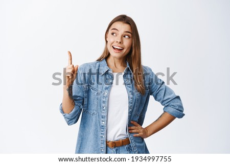 Thoughtful smiling woman with fair hair, raising finger and pointing up, having good point or idea, showing advertisement, standing against white background Royalty-Free Stock Photo #1934997755