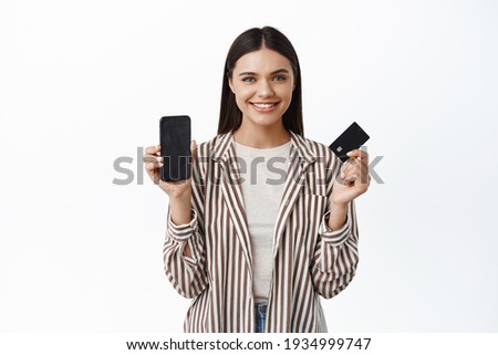 Portrait of modern young woman in stylish outfit, showing smartphone empty screen and plastic credit card, smiling pleased, recommending an app, white background