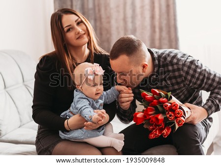 Young beautiful family with a little daughter on the couch with a bouquet of flowers. The father kisses the hand of the girl infant. Mother's Day