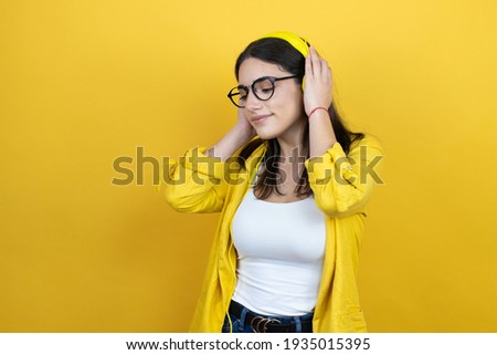 Young brunette businesswoman wearing yellow blazer over yellow background wearing headphones listening to music and dancing with a happy face standing and smiling with a confident smile showing teeth