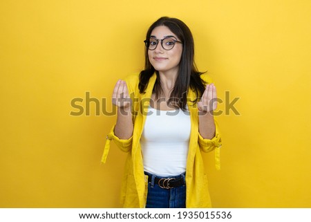Young brunette businesswoman wearing yellow blazer over yellow background doing money gesture with hands, asking for salary payment, millionaire business