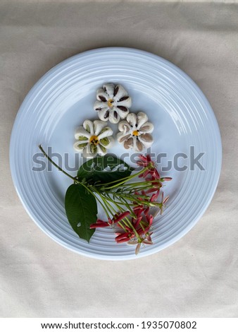 The picture of Thai dessert is called `”sunflower cookies mini”. It has several test delicious such as green tea, Black sesame seeds, Taro. It is on white plate and decorat  with red flower.