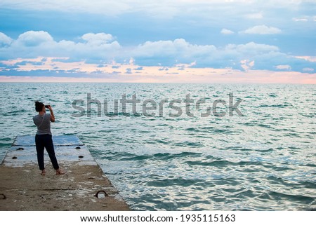 the girl stands on the beach on the pier in the summer and takes pictures and admires the beautiful sunset. Beautiful bluish-green water color and gray-blue clouds