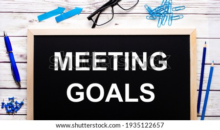 MEETING GOALS written on a black note-board next to blue paper clips, pencils and a pen.