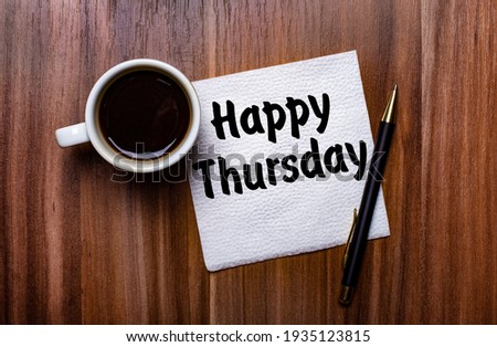 On a wooden table next to a white cup of coffee and a pen is a white paper napkin with the words HAPPY THURSDAY Royalty-Free Stock Photo #1935123815