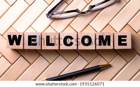 The word WELCOME is written on wooden cubes on a wooden background next to a pen and glasses.