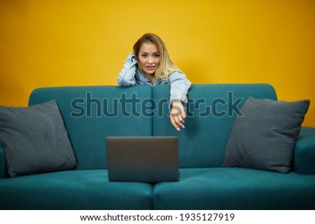 Colorful studio portrait of young attractive blonde girl using computer against yellow wall. Photo of happy young blonde woman standing isolated over yellow background sit on the bed