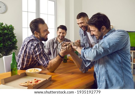 Four adult friends have fun together on the weekend. Cheerful company of male friends at home drinking beer and competing in arm wrestling. Concept of friendly competition, strength and men's leisure.