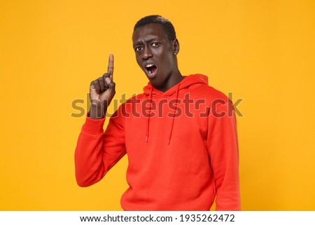 Shocked irritated young african american man guy in red streetwear hoodie posing isolated on yellow wall background. People lifestyle concept. Mock up copy space. Pointing index finger up, swearing