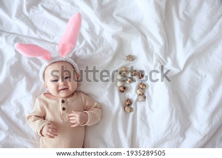 An infant dressed up bunny ears, smiling and looking at the camera, lying on the white sheet with small quail eggs. Easter baby as a rabbit. Spring hunting concept in a bunny costume.
