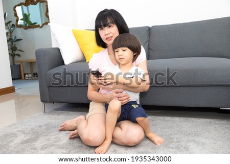 Asian Mom and son couple watching TV on a sofa at home, mom teaching son how to use remote control.