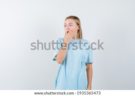  blonde girl in blue t-shirt covering mouth with hand, looking away and looking surprised, front view. 