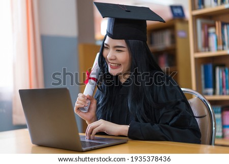 A young Asian university graduate woman in graduation gown expressing joy and excitement to celebrate her education achievement in front of a laptop making a remote video call to her parents at home