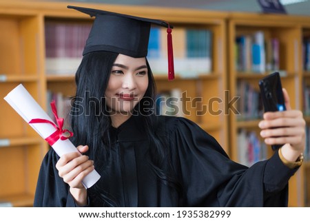 Young cheerful Asian university graduate woman in graduation gown and cap with a degree certificate in hand make a phone call to friends to celebrate her education achievement on the commencement day