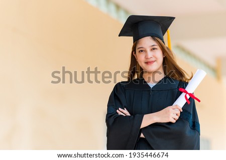Smiling female Asian student in academic gown and graduation cap holding diploma Royalty-Free Stock Photo #1935446674