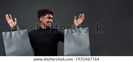Smiling young african american man holding bags after shopping and looking to side while posing on dark background, panorama. Discount and sale concept