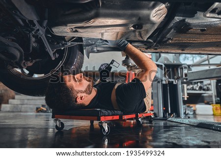 Male car mechanic worker working using wrench tool for repair, maintenance underneath car. Mechanic vehicle service checking under car in garage. Auto car repair service, maintenance concept. Royalty-Free Stock Photo #1935499324