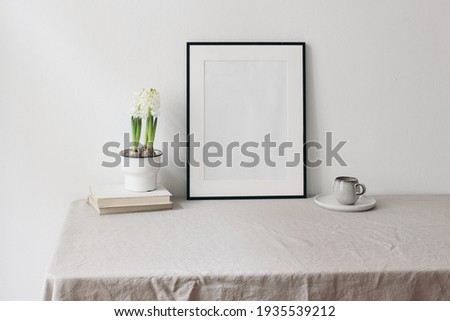 Easter spring still life. Blank black picture frame mockup on linen table cloth. Old books on cup of coffee and hite hyacinth in flower pot. Home office concept. Scandinavian interior design.