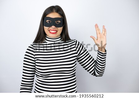 Young beautiful brunette burglar woman wearing mask doing hand symbol