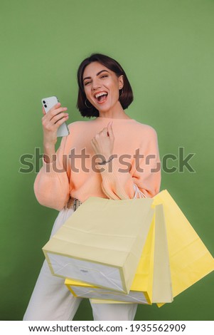 Young woman in casual peach sweater isolated on green olive color background doing winner gesture with mobile phone, holding shopping bags, discount sale concept