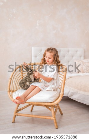little girl in a white dress is playing with a rabbit. A child in a bright room is sitting in a chair with a toy rabbit..