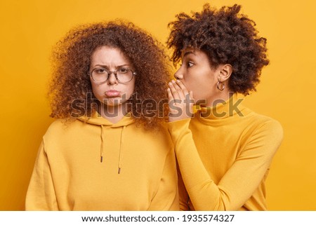 Surprised African American woman whispers secret information on ear of best friend who looks with gloomy expression spread rumors tells private news isolated over yellow background. Secrecy concept Royalty-Free Stock Photo #1935574327
