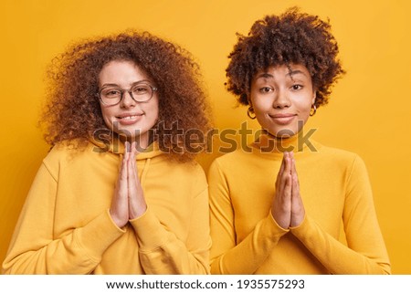 Hopeful diverse young women with curly hair plead for mercy look with beseech expressions keeps palms pressed together ask for help stand closely to each other isolated over yellow background