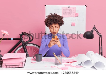 Photo of curly haired busy female student poses in coworking space works on blueprints prepares coursework searches information via smartphone connected to internet isolated on pink background