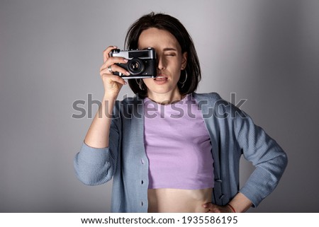 A young girl in a sweater and a top poses with a vintage film camera on a light background.