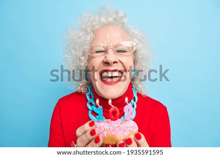 Happy birthday concept. Overjoyed senior lady smiles broadly has white perfect teeth going to blow candles on glazed doughnuts being well dressed celebrates 102nd bday enjoys party with relatives