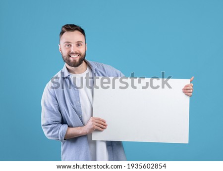 Joyful Caucasian man demonstrating white blank placard on blue studio background, mockup for design