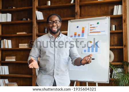 African American school teacher in glasses standing next to whiteboard with bar charts, explaining mathematics, streaming lesson online and looking at webcam, teaching in a virtual online classroom Royalty-Free Stock Photo #1935604321