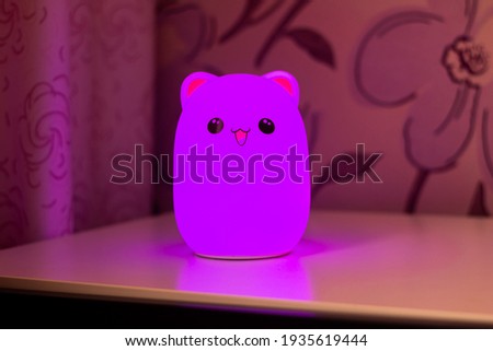The baby cute bear-shaped purple night lamp with eyes and ears on the bedside table in the pink-style room glows in the dark