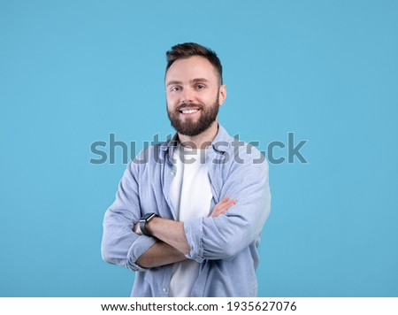 Portrait of positive young guy posing with crossed arms and smiling at camera on blue studio background Royalty-Free Stock Photo #1935627076