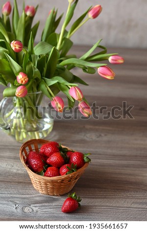A basket of ripe strawberries and a jug of tulips on a wooden table. Image with selective focus.
