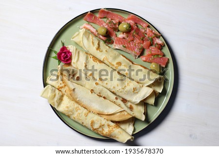 Pancakes on green plate with salty salmon slices, olives, rose flower on white table