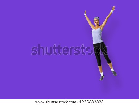 Full body of happy excited woman jumping or doing fitness exercise, isolated over violet purple color background. Fit girl with raised up hands, in sportswear, at studio.