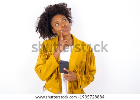 young beautiful African American woman wearing yellow jacket over white wall thinks deeply about something, uses modern mobile phone, tries to made up good message, keeps index finger near lips.